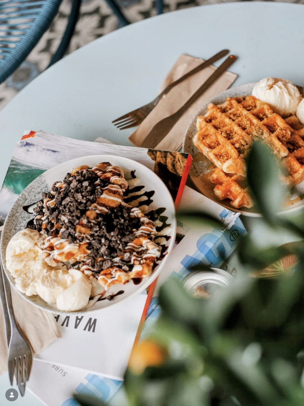 Chocolate-drizzled acai bowl with ice cream and waffles served on a pale-blue table next to a Saltd surf-illustrated menu booklet