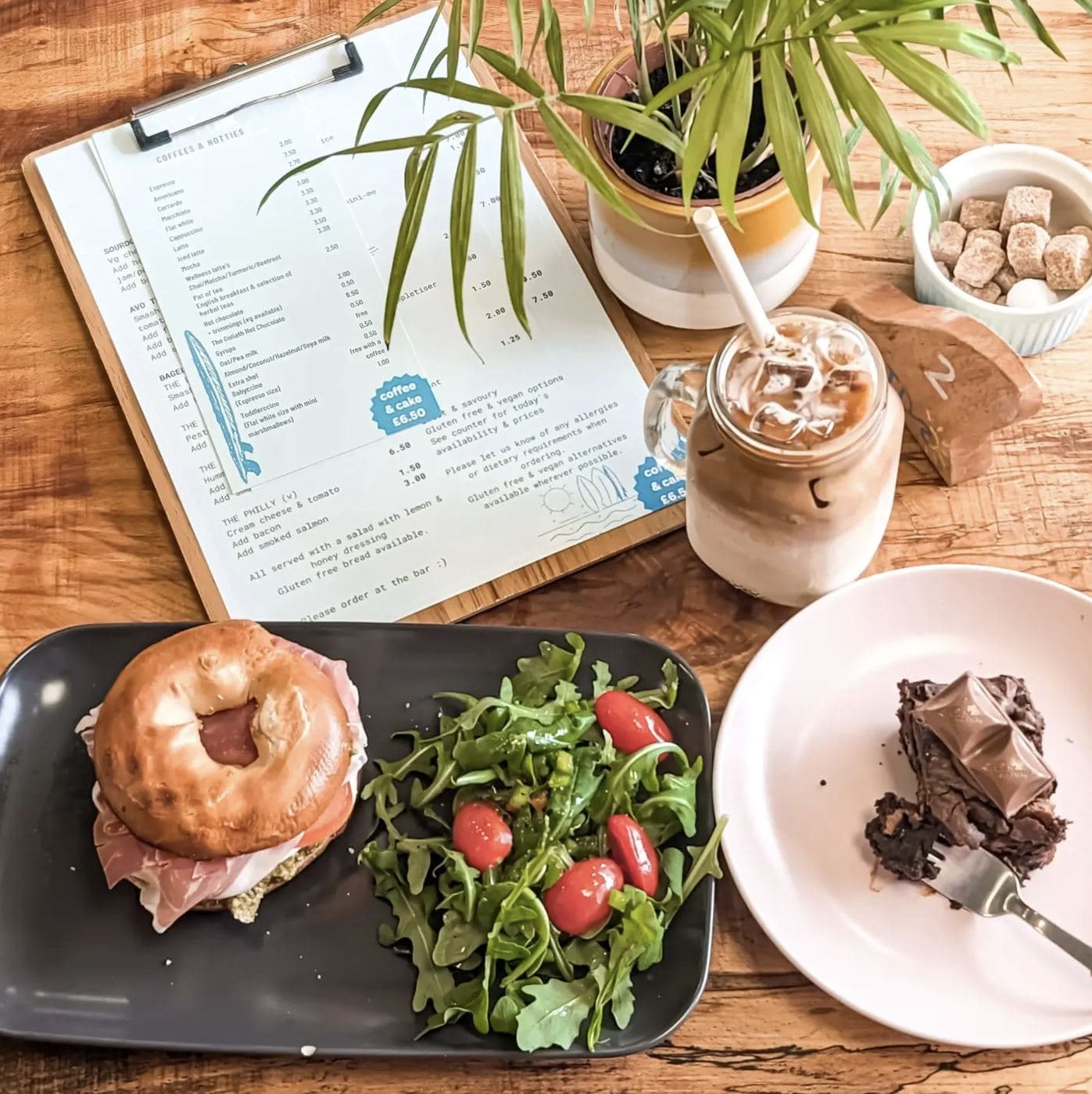 A Saltd lunch spread — bagel with prosciutto, salad with rocket and tomato, an iced coffee and a chocolate brownie alongside the open Saltd coffee-and-bakes menu