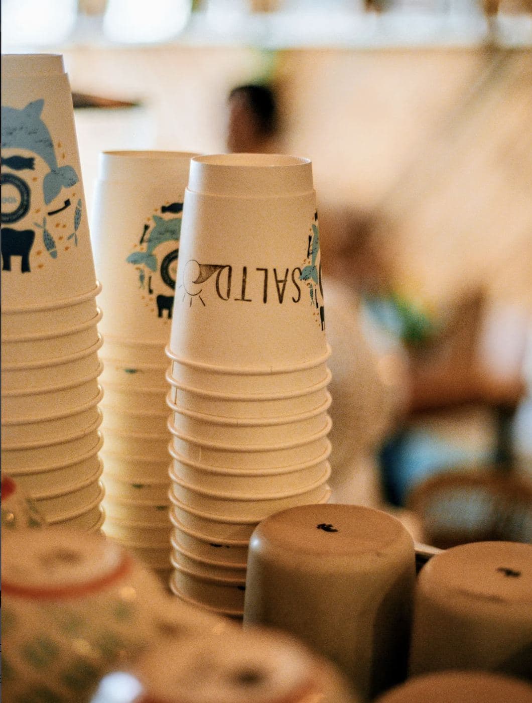 Stacked SALTD-branded paper coffee cups showing the Saltd wordmark with the wave-and-sun logo, ready behind the counter