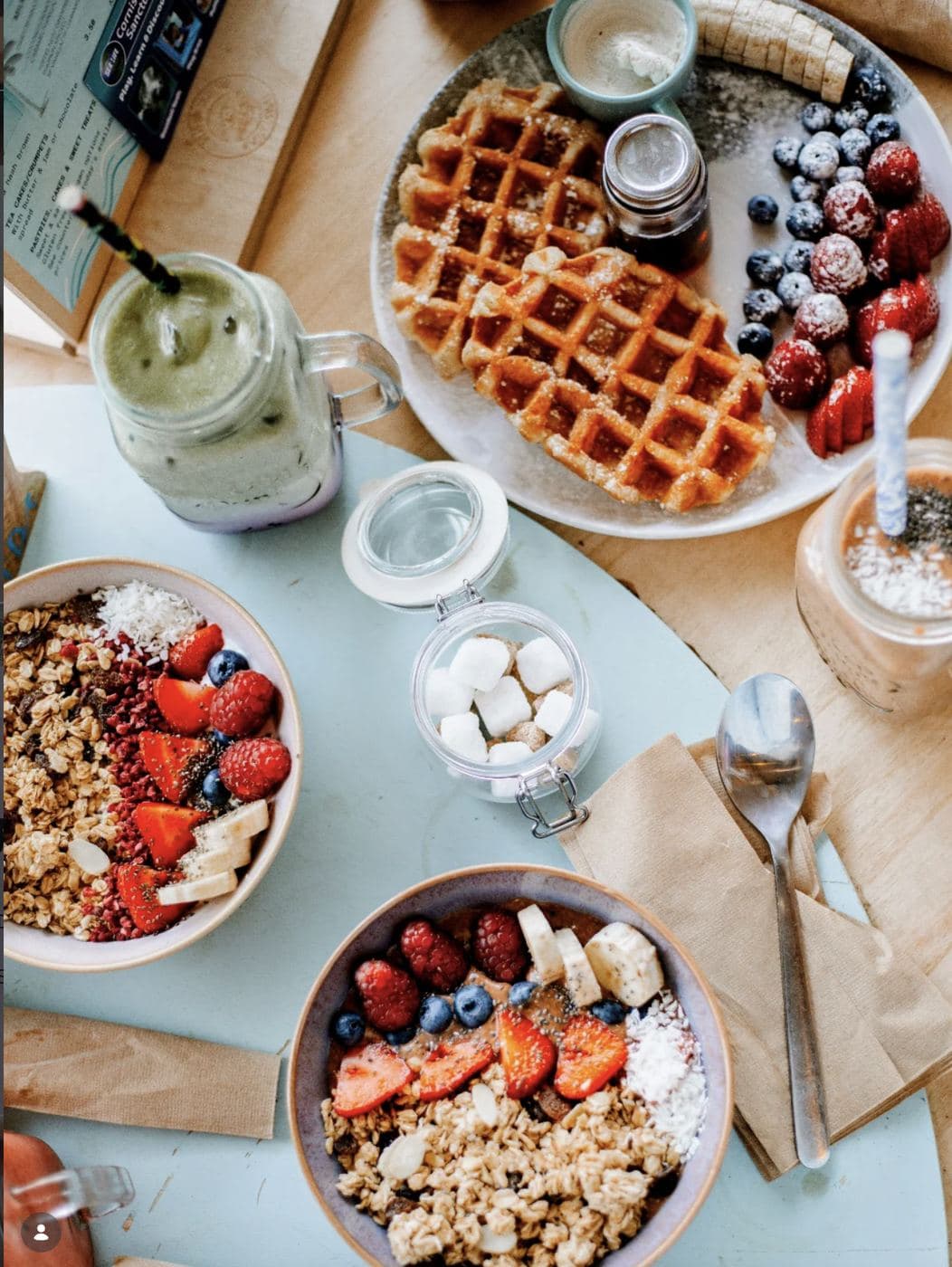 Saltd brunch spread overhead — two acai bowls topped with fruit, waffles with berries and banana, a green matcha smoothie and an iced coffee on a wooden table