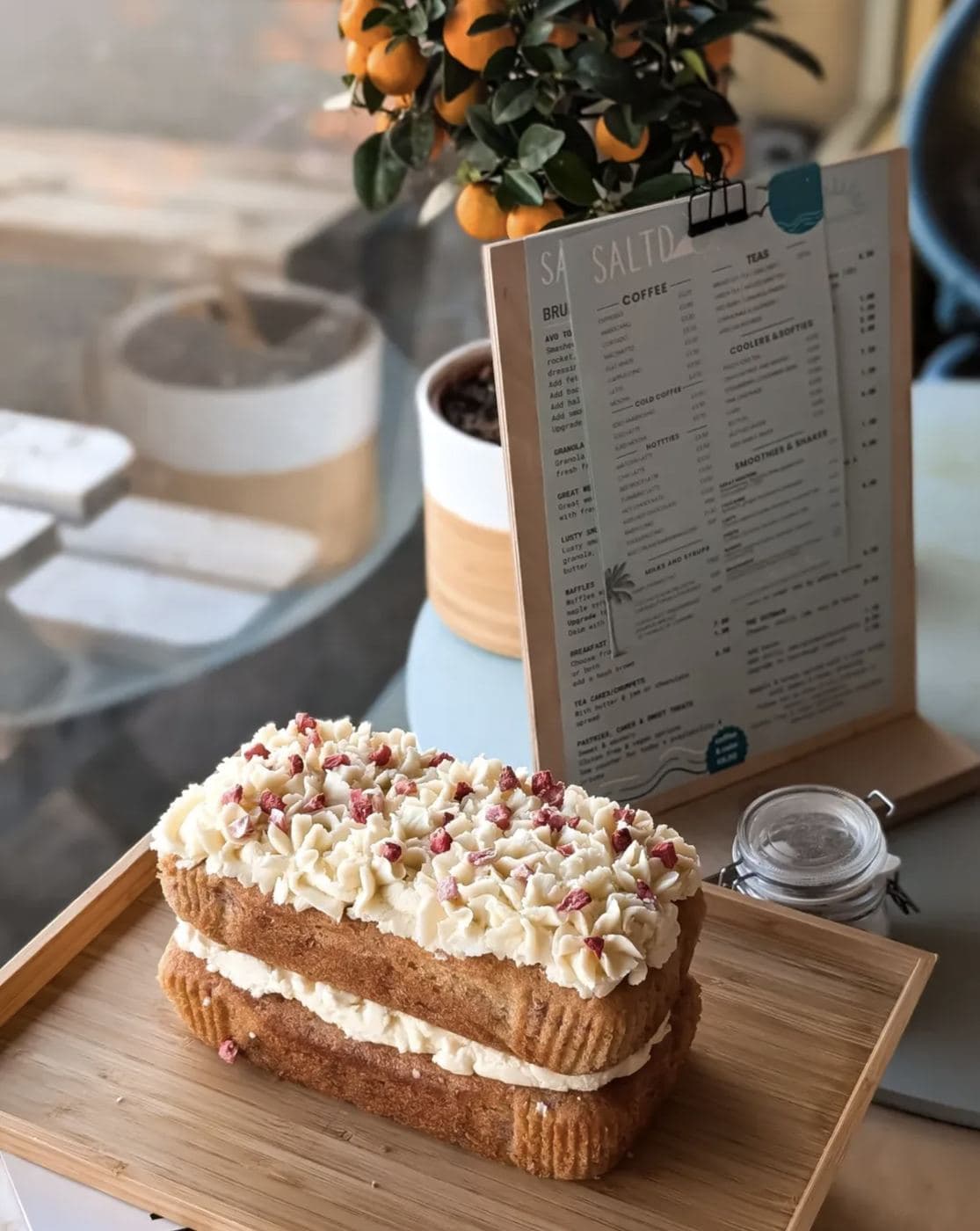 A pomegranate and pistachio layered cake on a wooden board, photographed in front of the standing Saltd menu