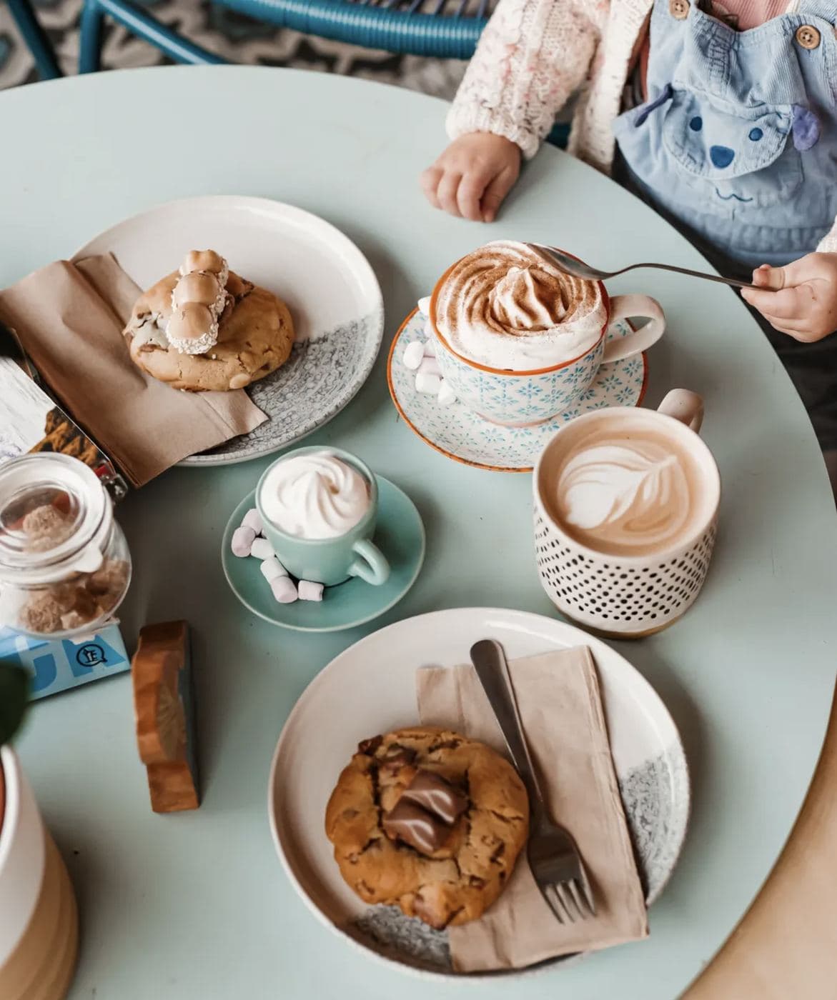Three Saltd coffees on a pale-blue table — cappuccino with art, leaf-foam latte and hot chocolate with marshmallows — alongside chocolate cookies