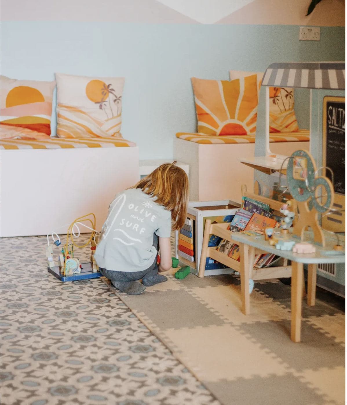 The Saltd kids' play area — a child in a 'Saltd Surf' t-shirt at the books-and-toys nook, with sunset-print pillows on benches behind