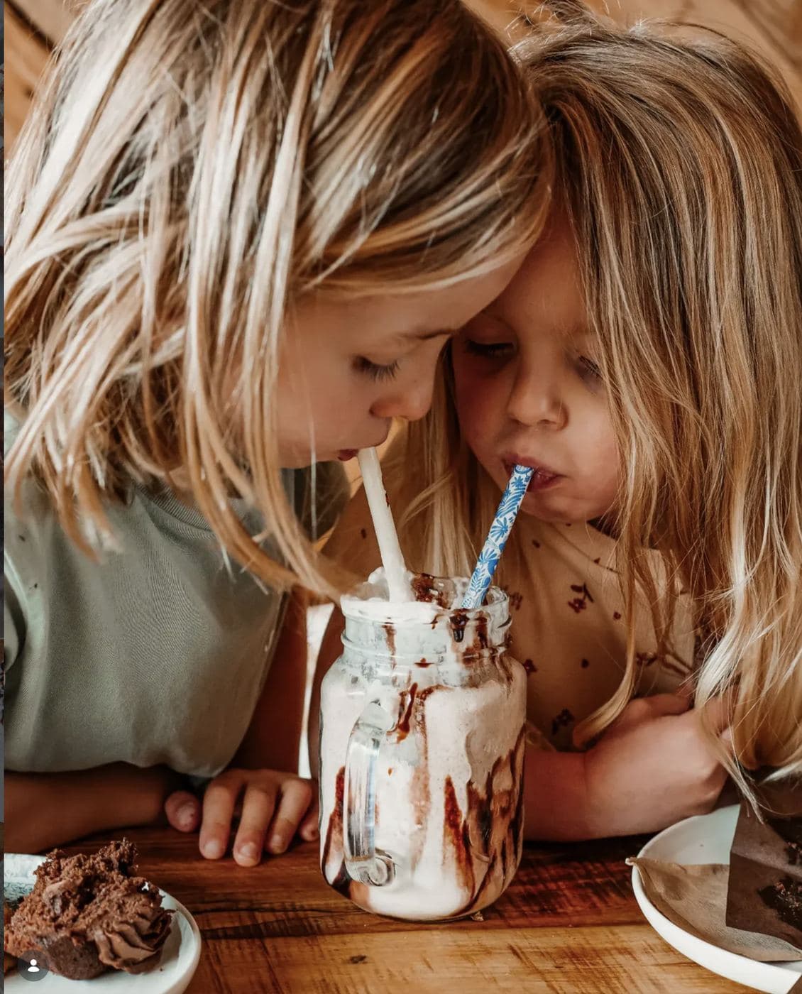 Two children leaning into a shared chocolate milkshake at a Saltd table, sipping from two patterned straws