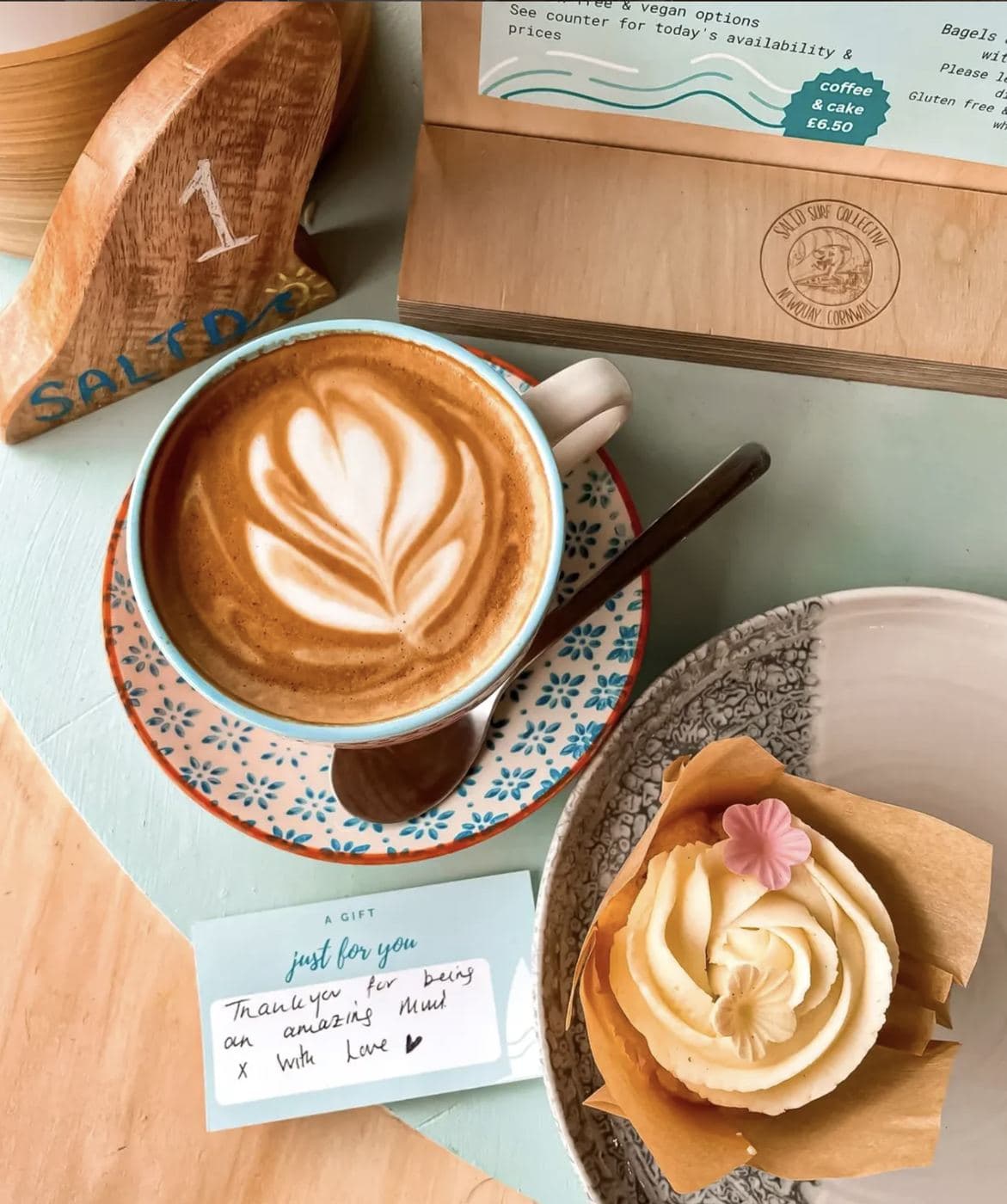 A Saltd latte with leaf foam art on a patterned saucer, served alongside a vanilla cupcake and a 'Thank you for being an amazing Mum' card