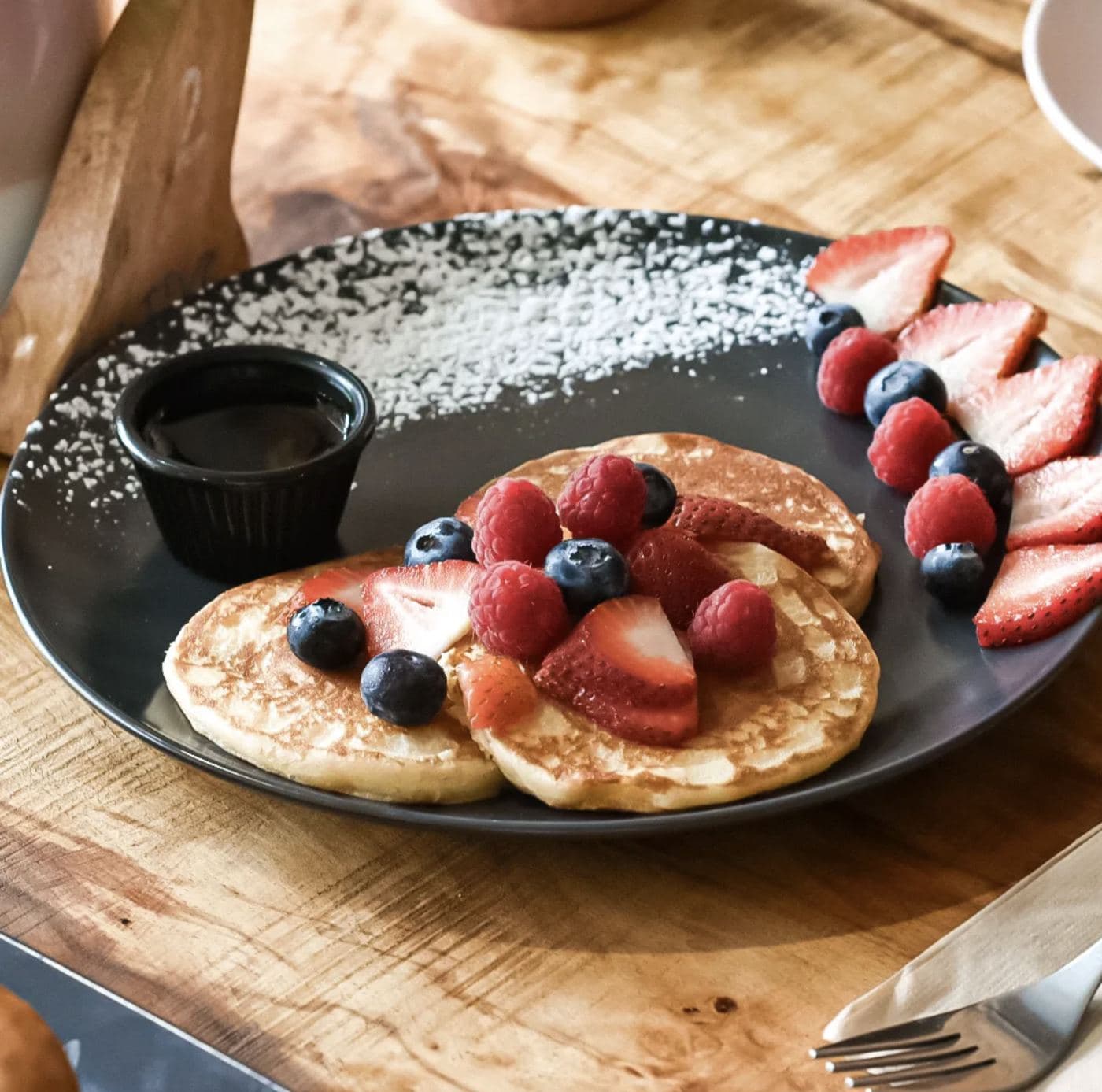 A stack of Saltd pancakes topped with raspberries, blueberries and sliced strawberries, served with a maple syrup pot and dusted with icing sugar on a wooden board